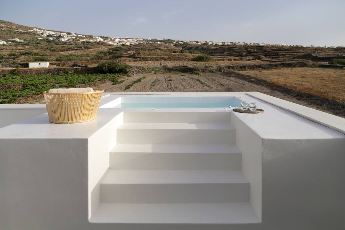 Una banyera hidromassatge moderna a l'aire lliure, amb uns escalons que baixen cap a l'aigua, envoltada per una estructura blanca i una vista de camps de conreu en terrasses al fons.