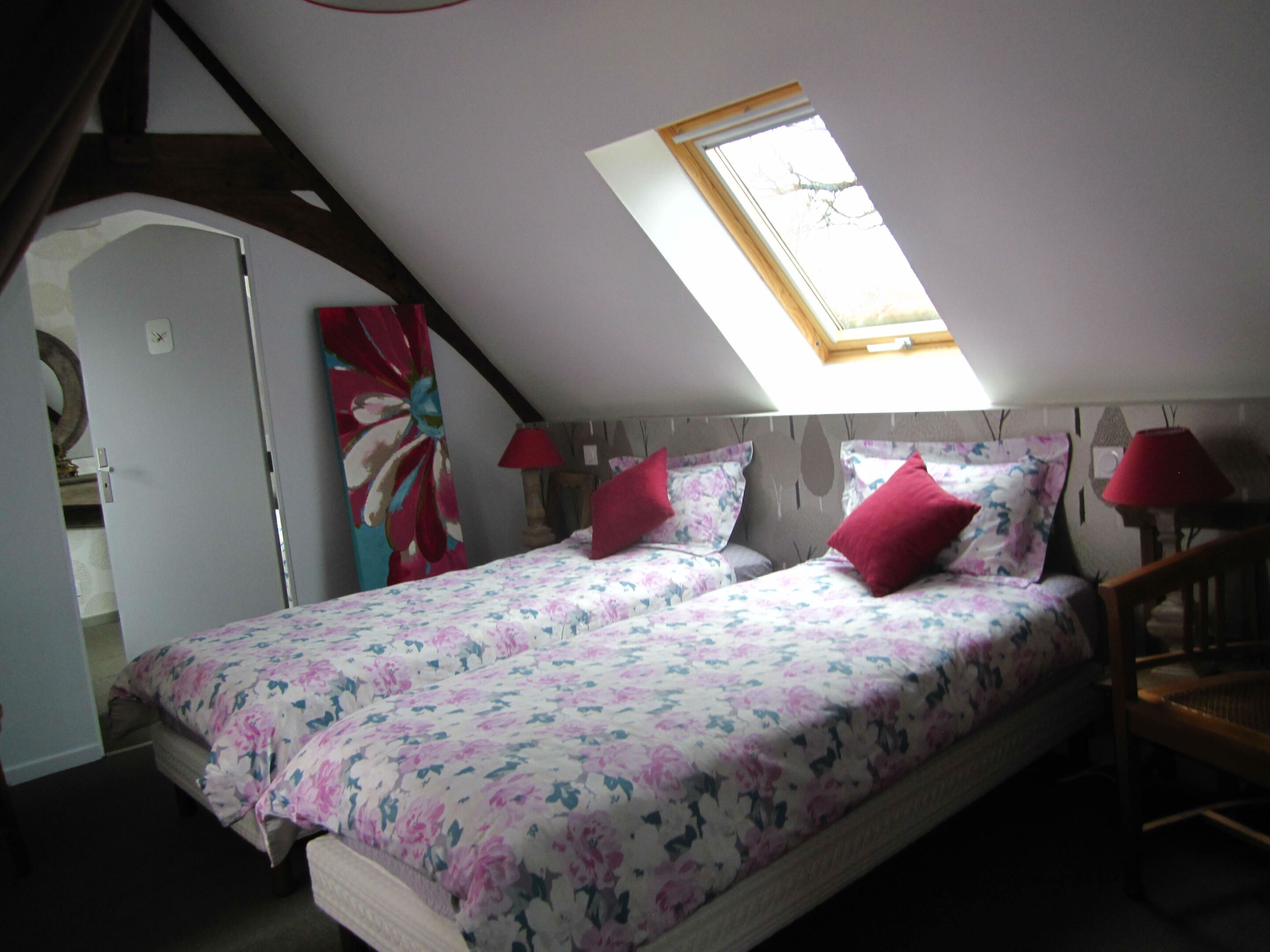 A bedroom with a sloped ceiling, a skylight, a bed with floral bedding, two red lamps, and a wooden chair.