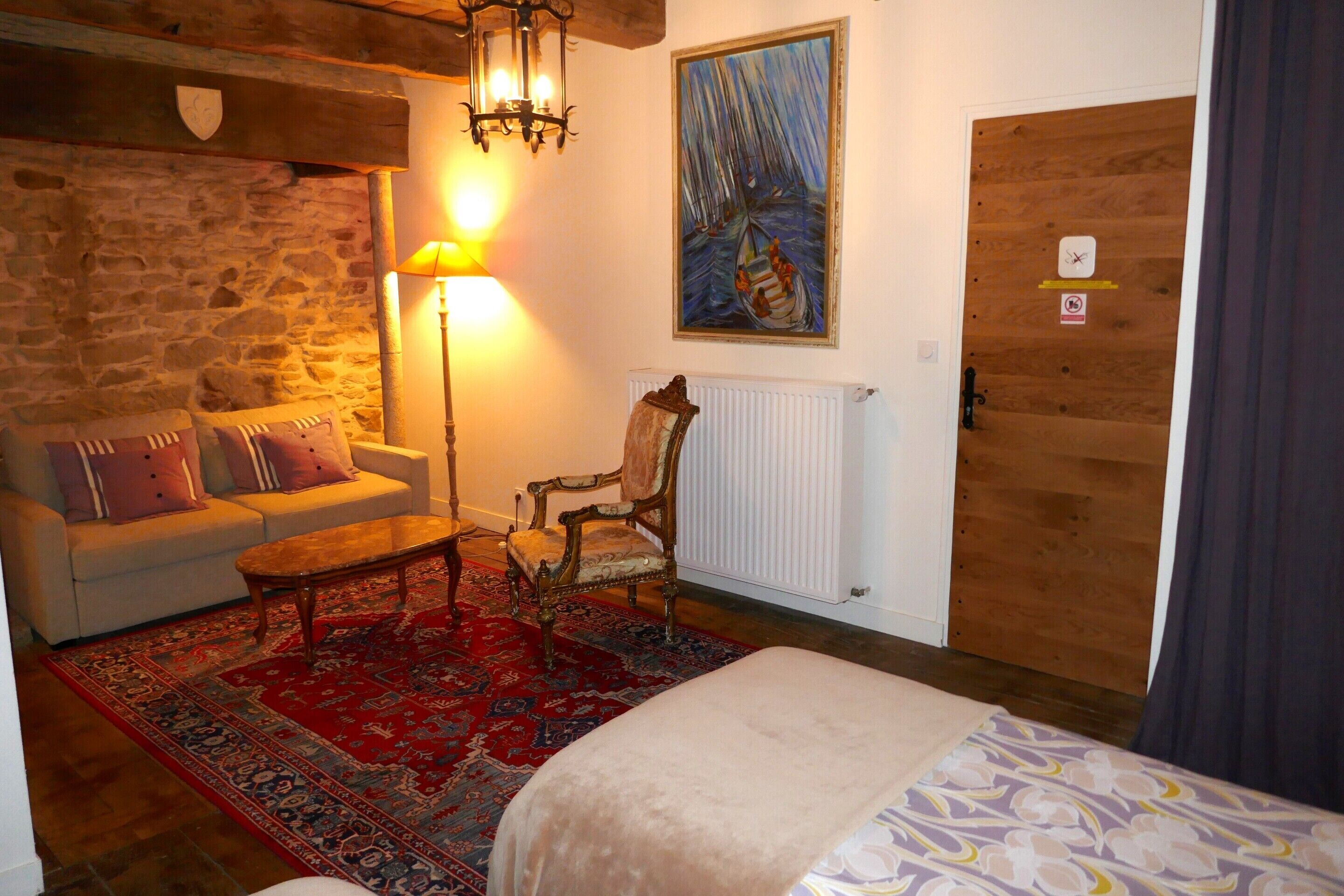 A cozy room with a stone wall, a wooden door, a painting, a sofa with cushions, a coffee table, and a bed with a patterned bedspread.