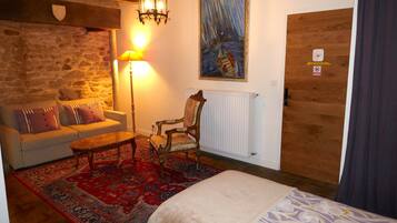 A cozy room with a stone wall, a wooden door, a painting, a sofa with cushions, a coffee table, and a bed with a patterned bedspread.