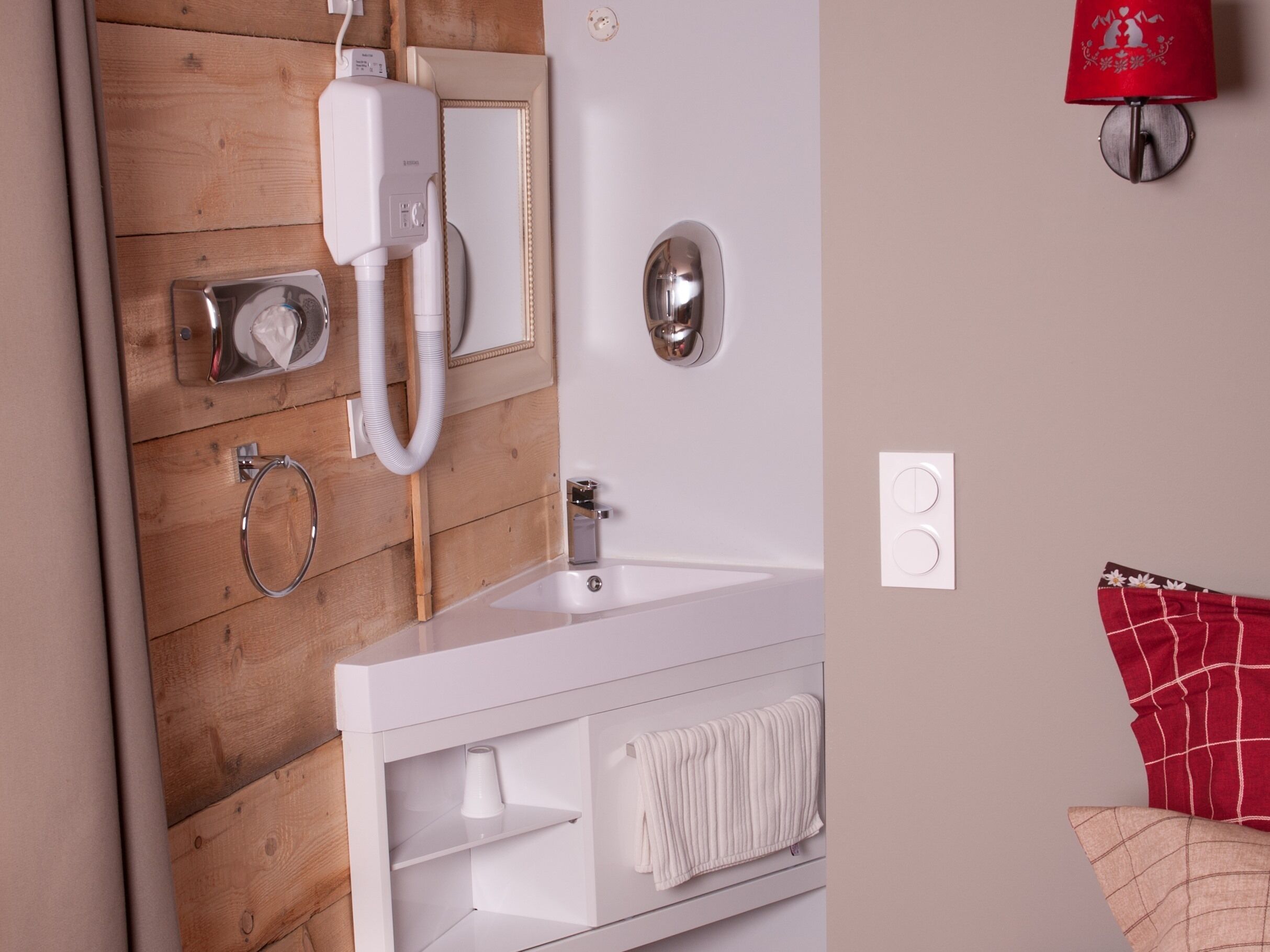 A modern bathroom with a wooden panel wall, a white sink, a mirror, a red wall-mounted light, and a towel rack.