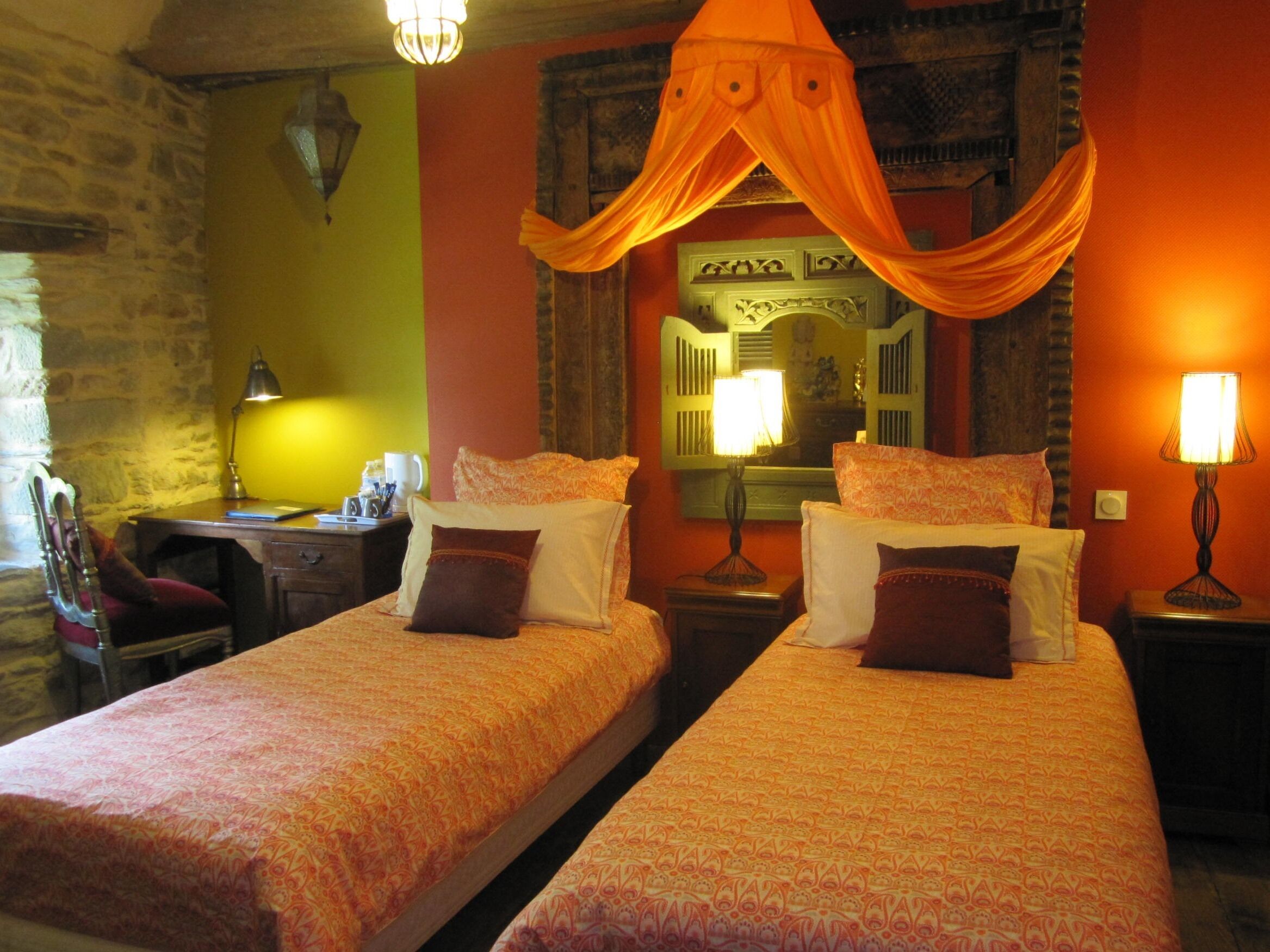 A room with two beds, each with patterned bedding and pillows, a desk with a lamp, and a chair.