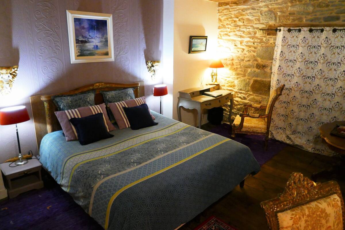 Une chambre à coucher avec un lit, deux tables de chevet, une commode, une chaise, une lampe et un mur en pierre.