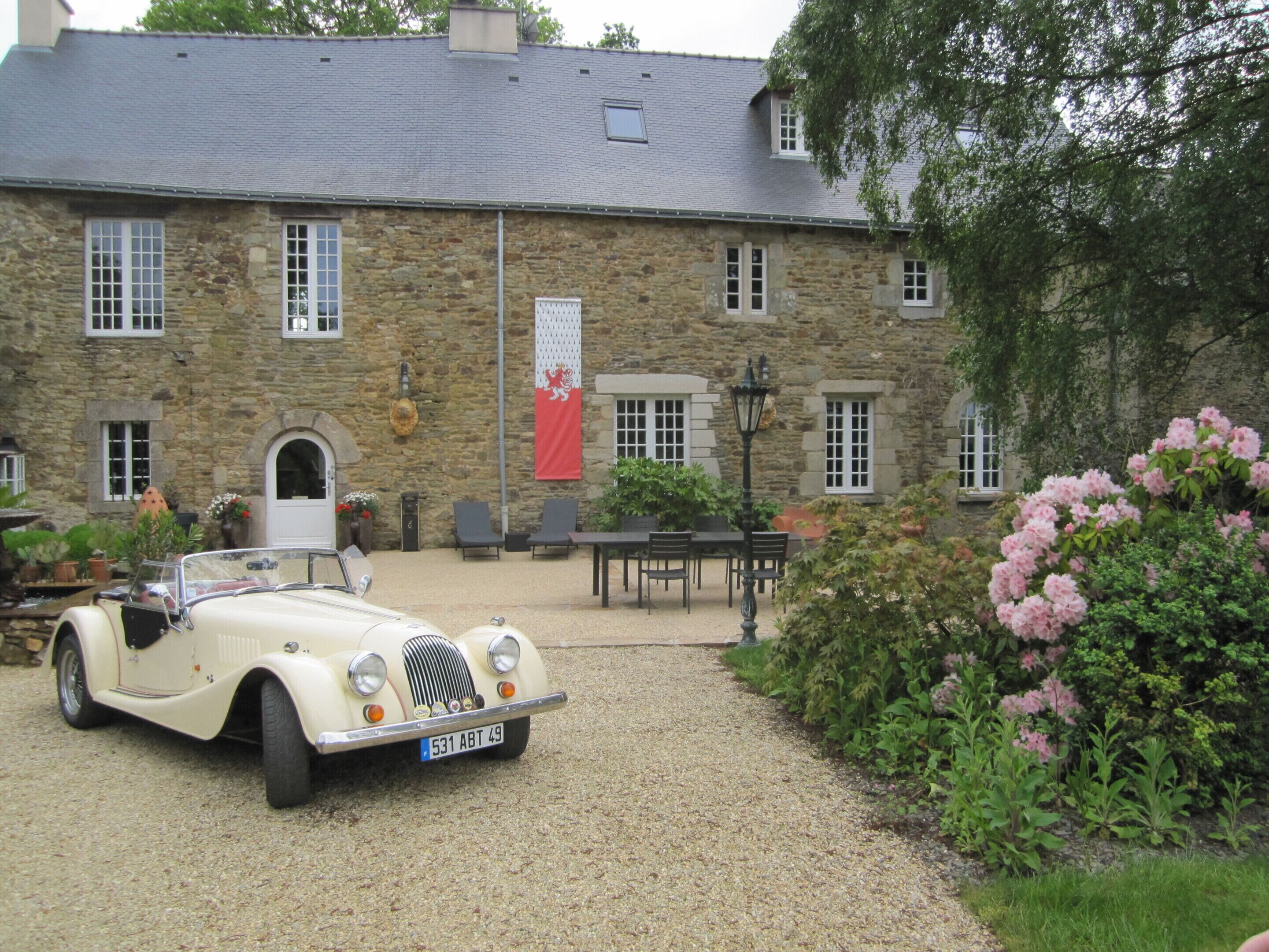 A vintage car parked in front of a stone building with a flag, outdoor seating, and a garden.
