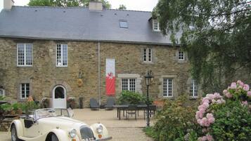 A vintage car parked in front of a stone building with a flag, outdoor seating, and a garden.