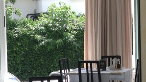Un coin repas avec une table dressée pour deux personnes, des chaises et une vue sur une haie verdoyante à l’extérieur.