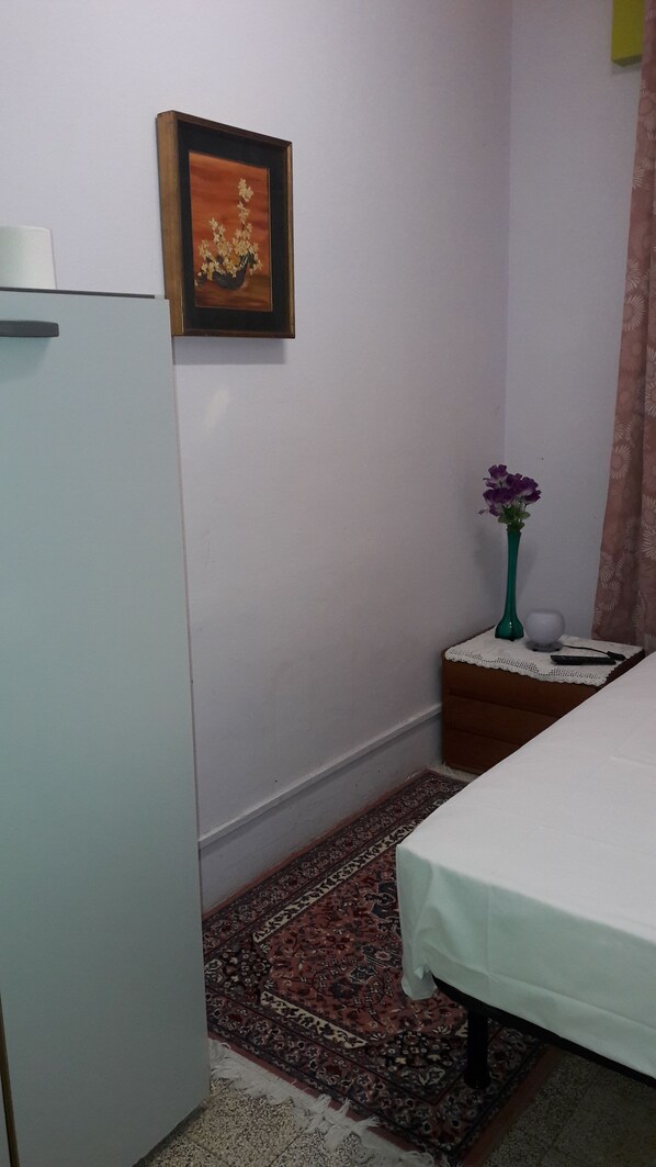 Une petite pièce avec un lit, une table de chevet ornée d’un vase de fleurs, un tableau encadré au mur et un tapis à motifs.