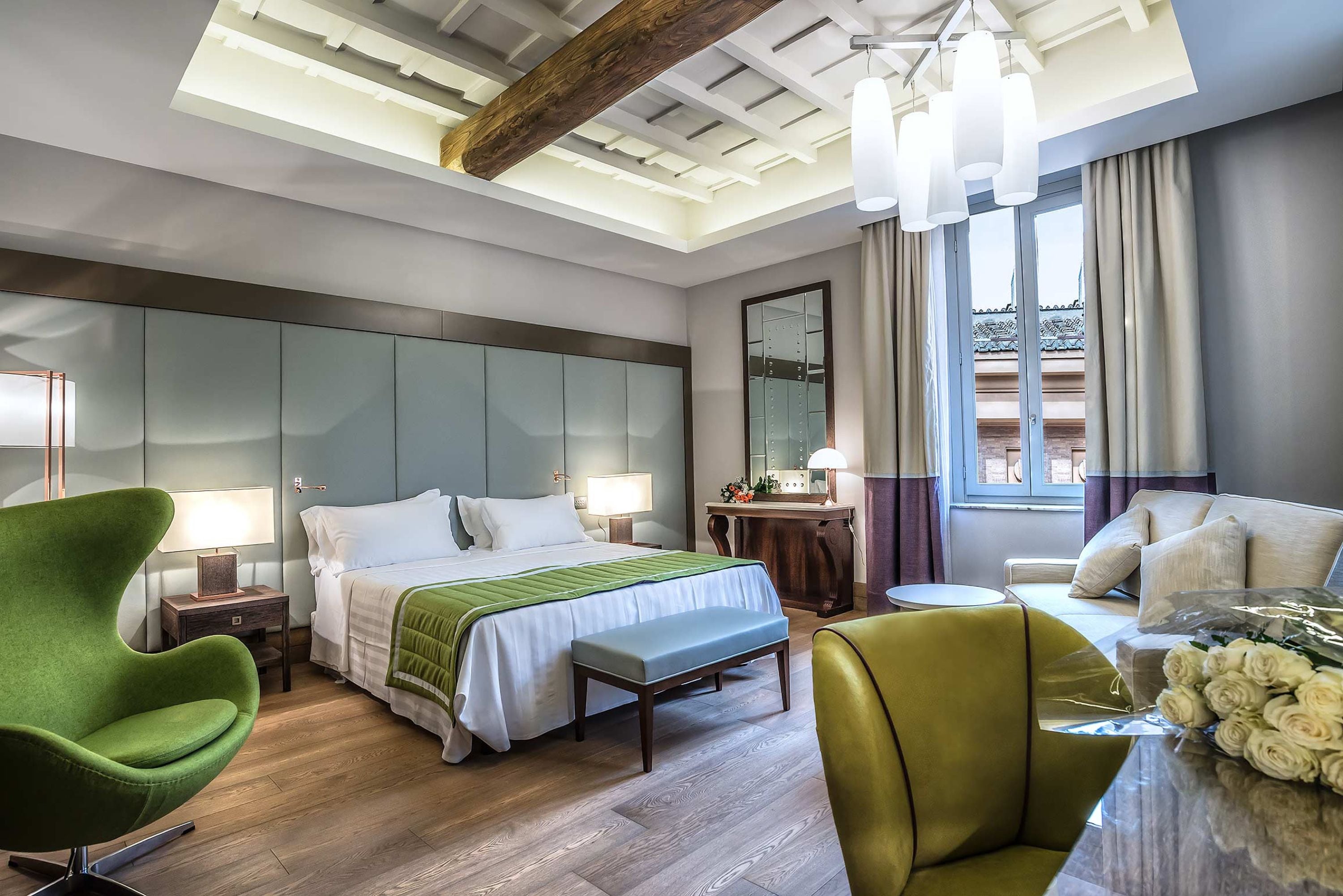 Habitación de hotel moderna con una cama grande, un sillón verde, un banco de madera, una pequeña mesa redonda con un florero y vistas al exterior a través de una ventana con cortinas.