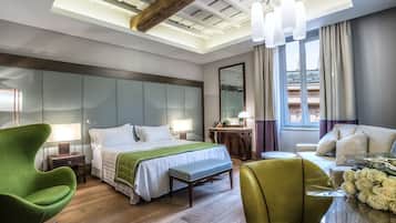 Habitación de hotel moderna con una cama grande, un sillón verde, un banco de madera, una pequeña mesa redonda con un florero y vistas al exterior a través de una ventana con cortinas.