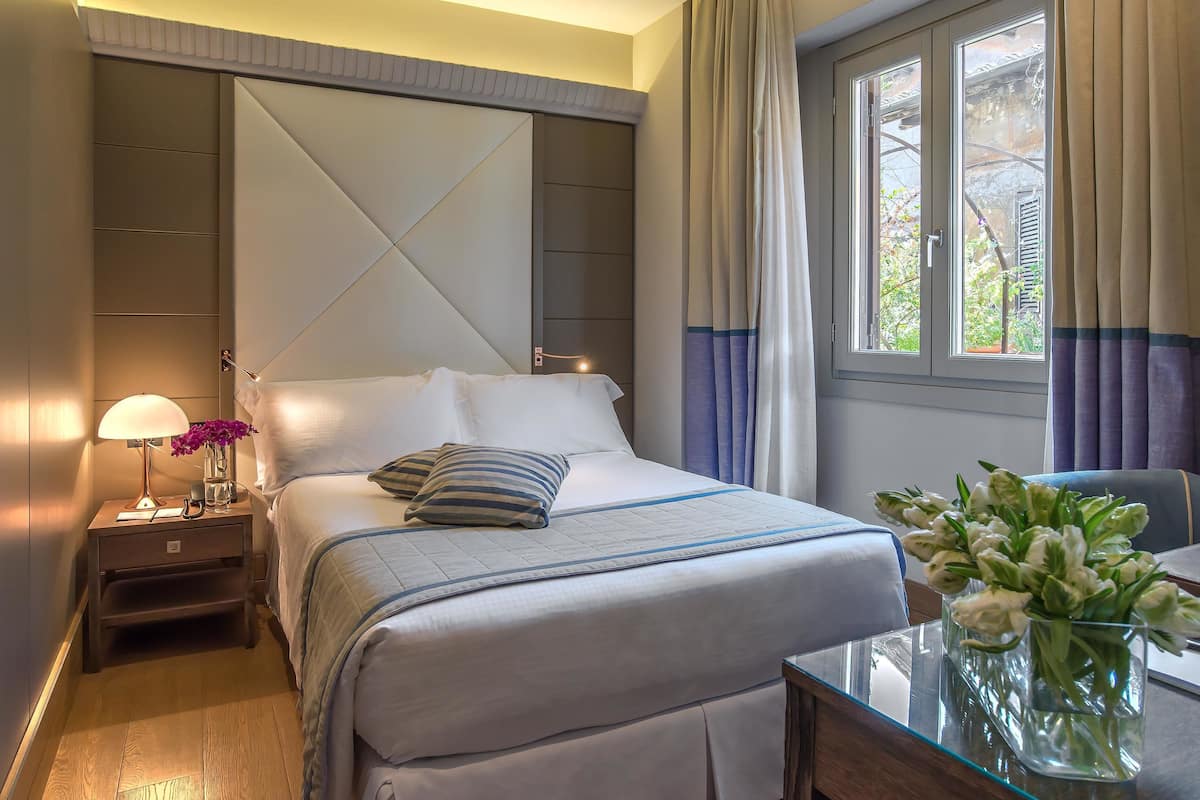 A neatly made bed with a striped pillow, a bedside table with a lamp and flowers, and a window with curtains.