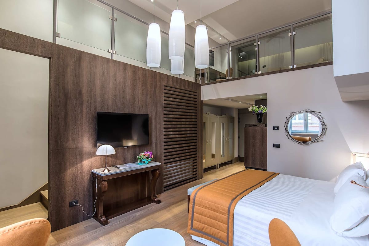 A modern hotel room with a large bed, a flat-screen TV, a desk with a lamp, and a balcony with glass railings.