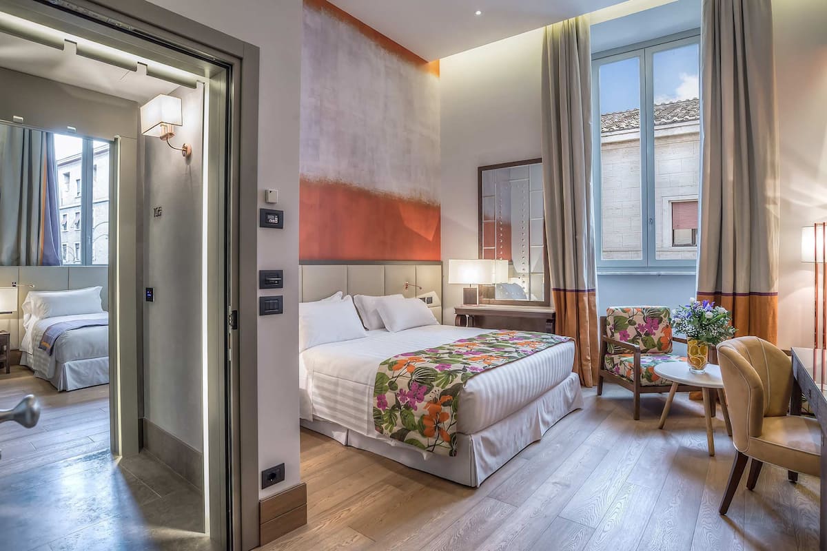 A hotel room with a bed, a desk, a chair, a window, and a door.