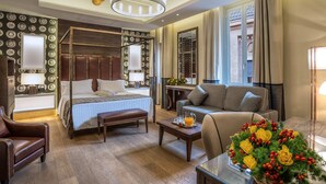 A hotel room with a four-poster bed, a sitting area with a sofa and armchair, a coffee table with a floral arrangement, and a dining table with a lamp. - Martis Palace Hotel (Rome)