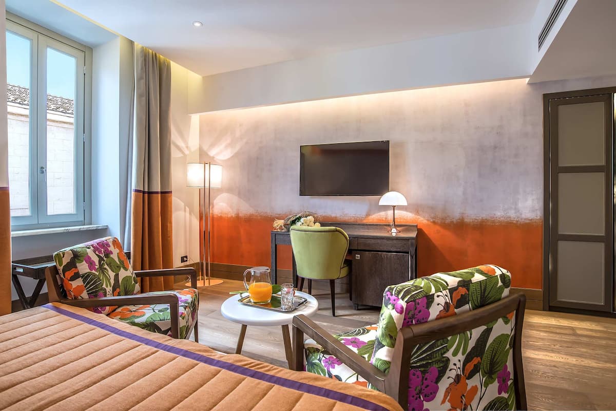 A hotel room with a bed, two chairs, a desk, a TV, and a small table with a tray.