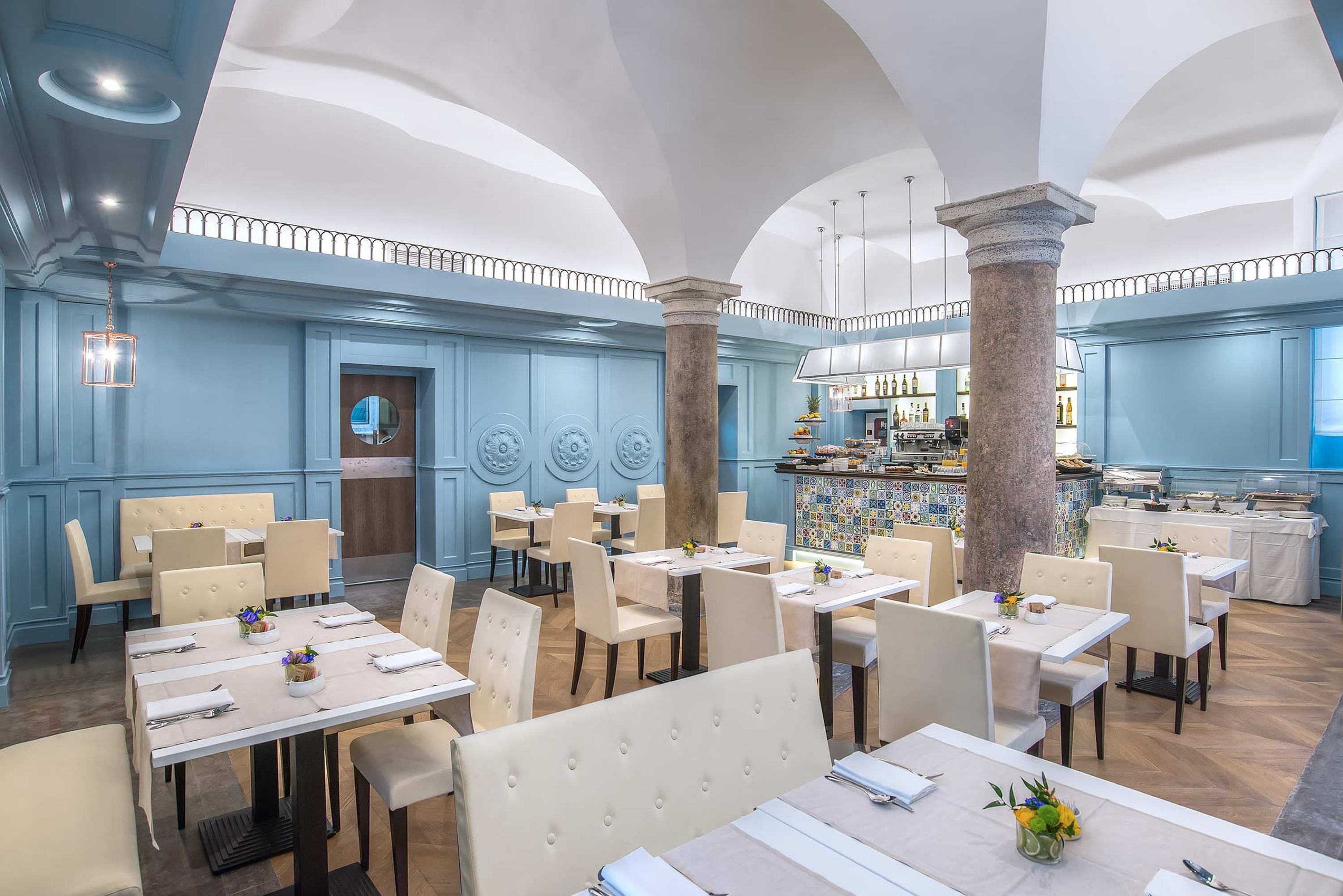 Ein Restaurant mit hohen Decken, blauen Wänden und einer Bar. Die Tische sind mit weißen Tischdecken und Stühlen gedeckt.