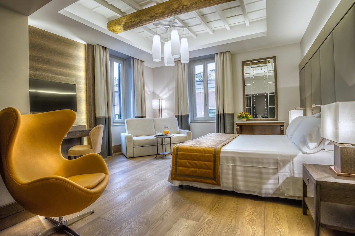 A modern hotel room with a large bed, a sofa, a unique yellow chair, a small table with a lamp, and a large mirror.