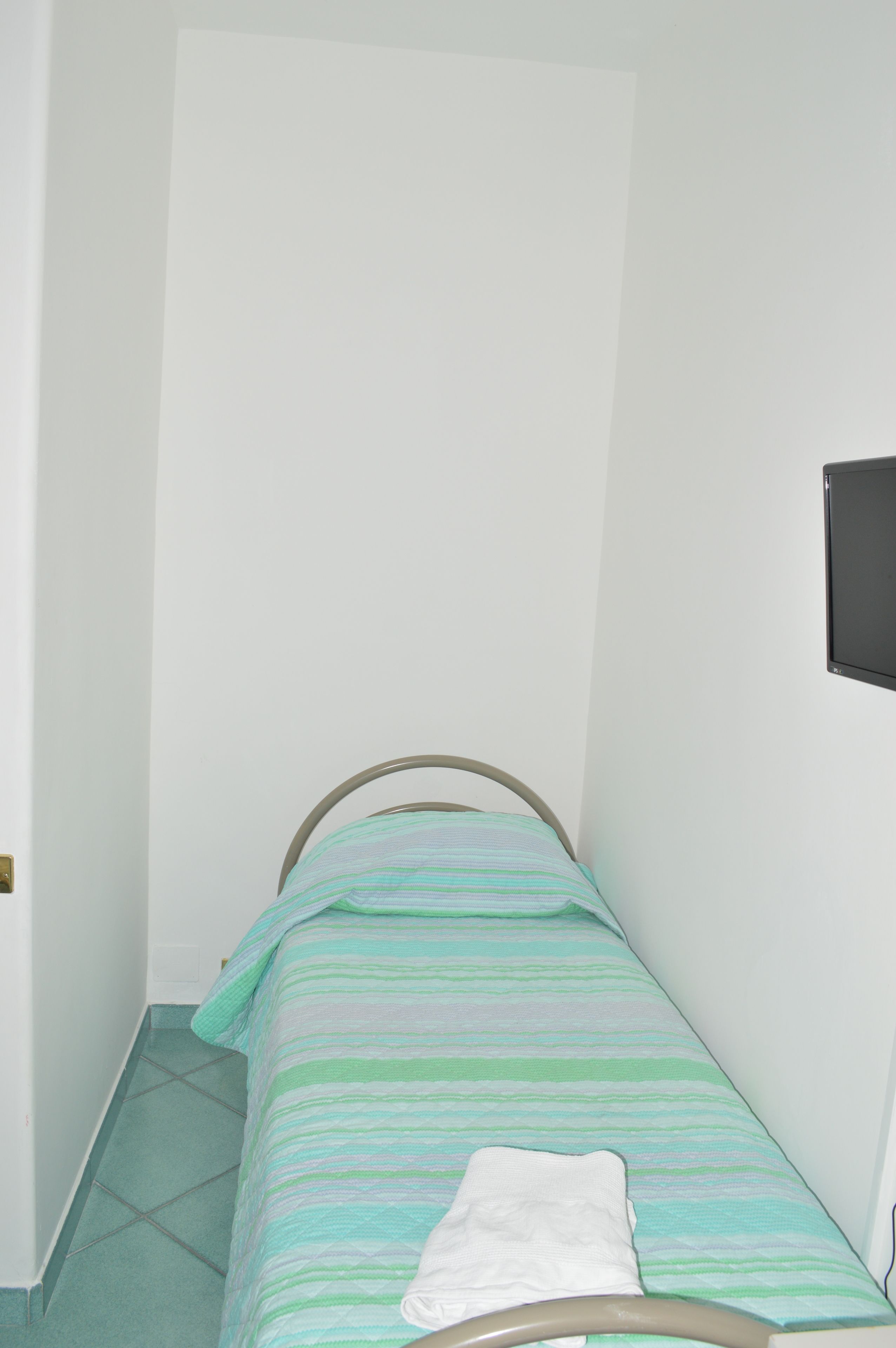 A small, narrow room with a single bed, a white wall, and a tiled floor.