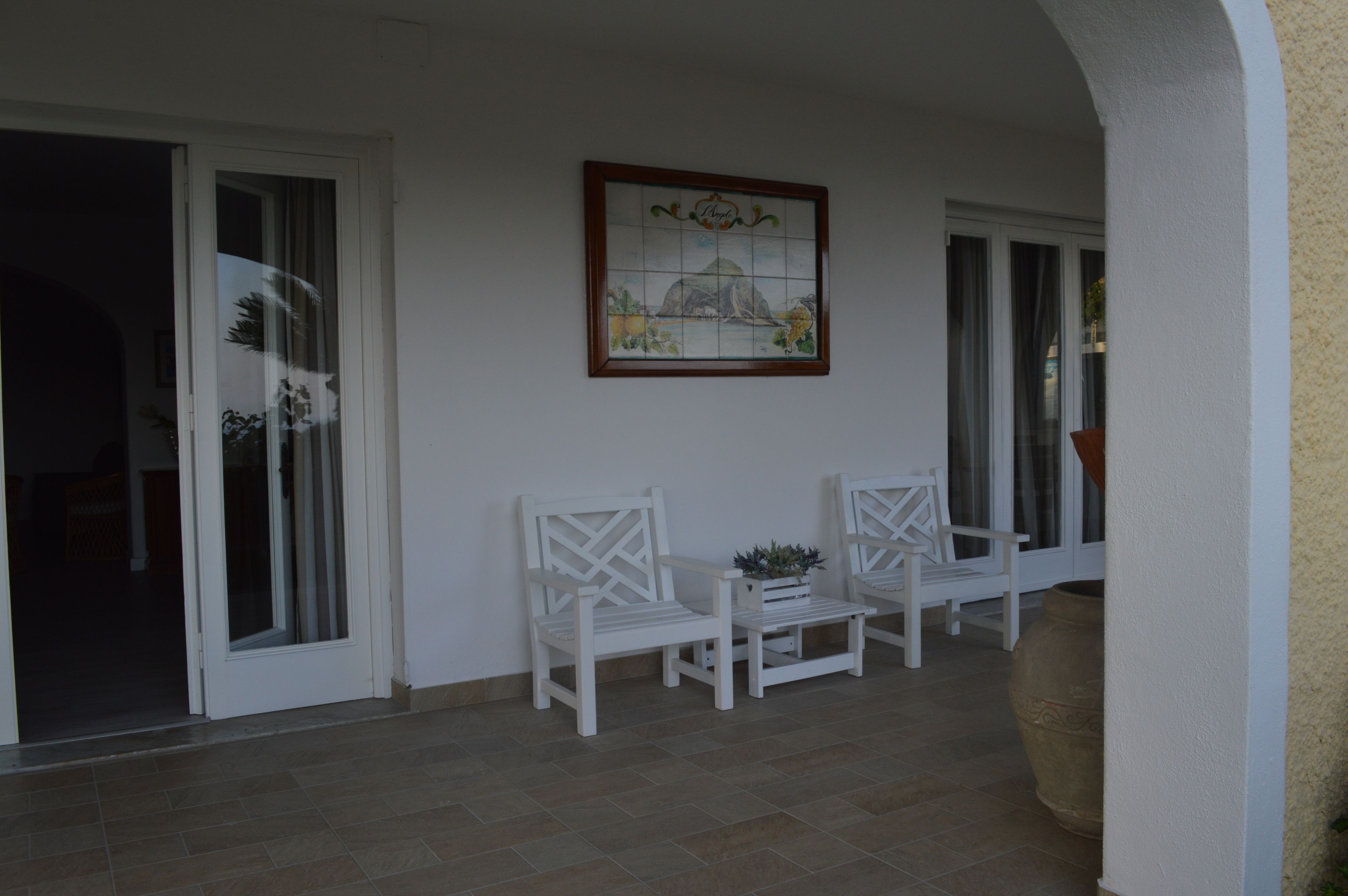 A covered outdoor seating area with two white chairs and a table, a large potted plant, and a framed artwork on the wall.