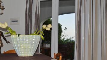 A dining table with a vase of white flowers, a wooden chair, and a view through a glass door to an outdoor area with greenery.