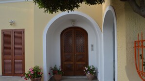 Entrada de un edificio con una puerta de madera grande, dos macetas y una pared blanca con un arco decorativo.