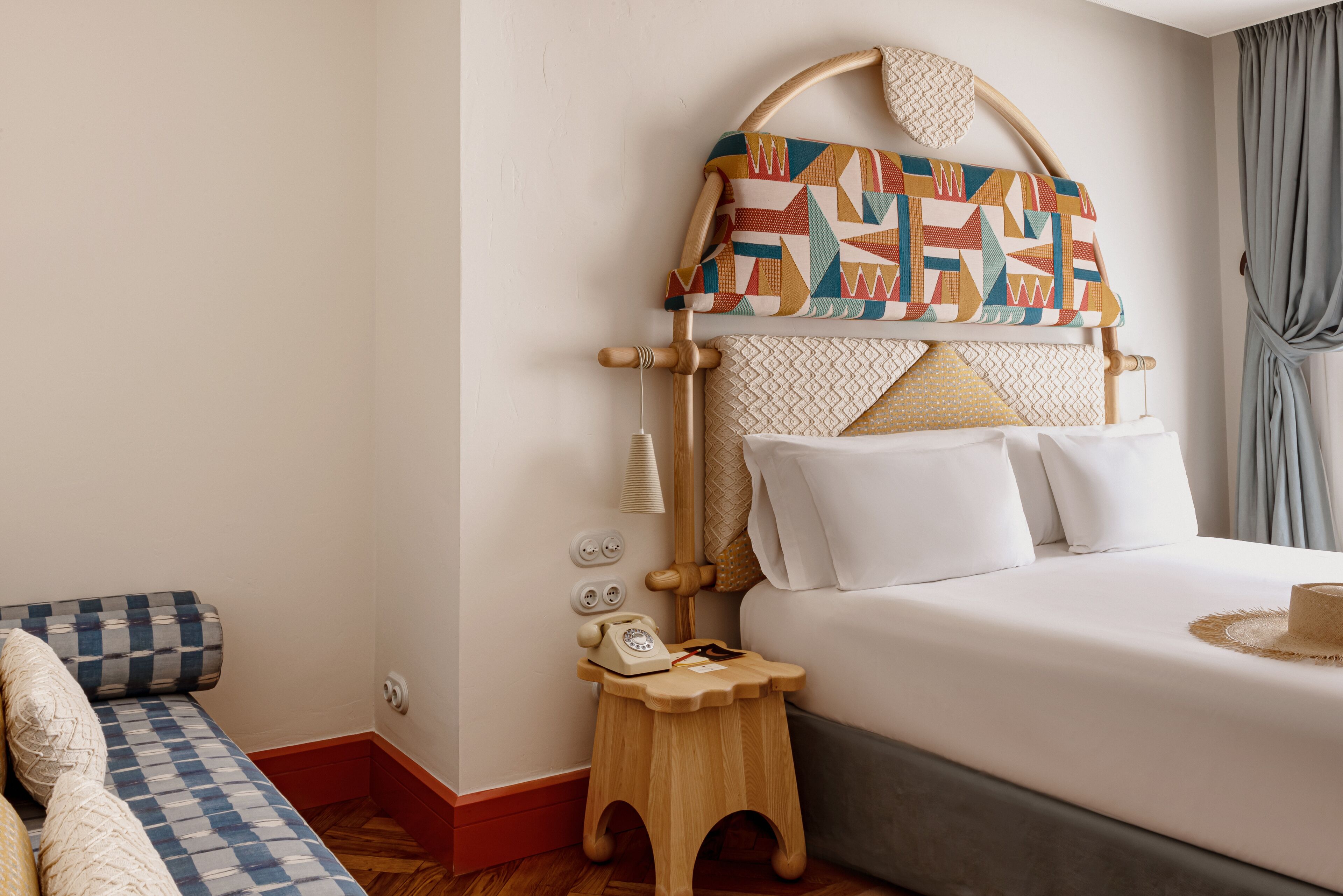 Ein ordentlich bezogenes Bett mit einem gemusterten Kopfteil, ein Nachttisch mit Telefon und eine karierte Bank in einem Hotelzimmer.