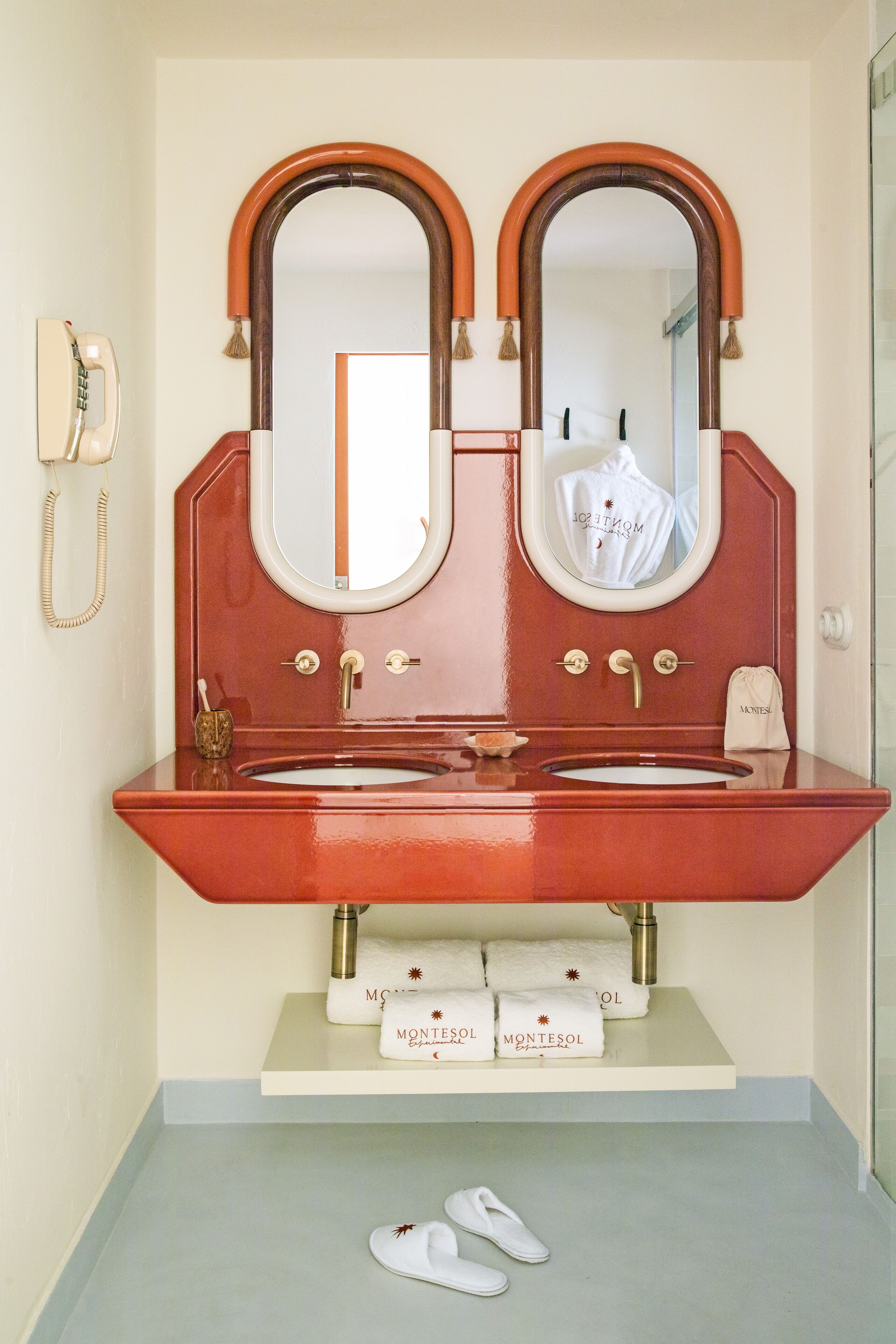 Un baño con un lavamanos rojo, dos espejos, un teléfono y un estante con toallas.