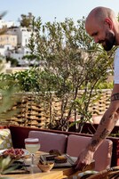 Un hombre sirve comida en una mesa al aire libre, con edificios y árboles de fondo.