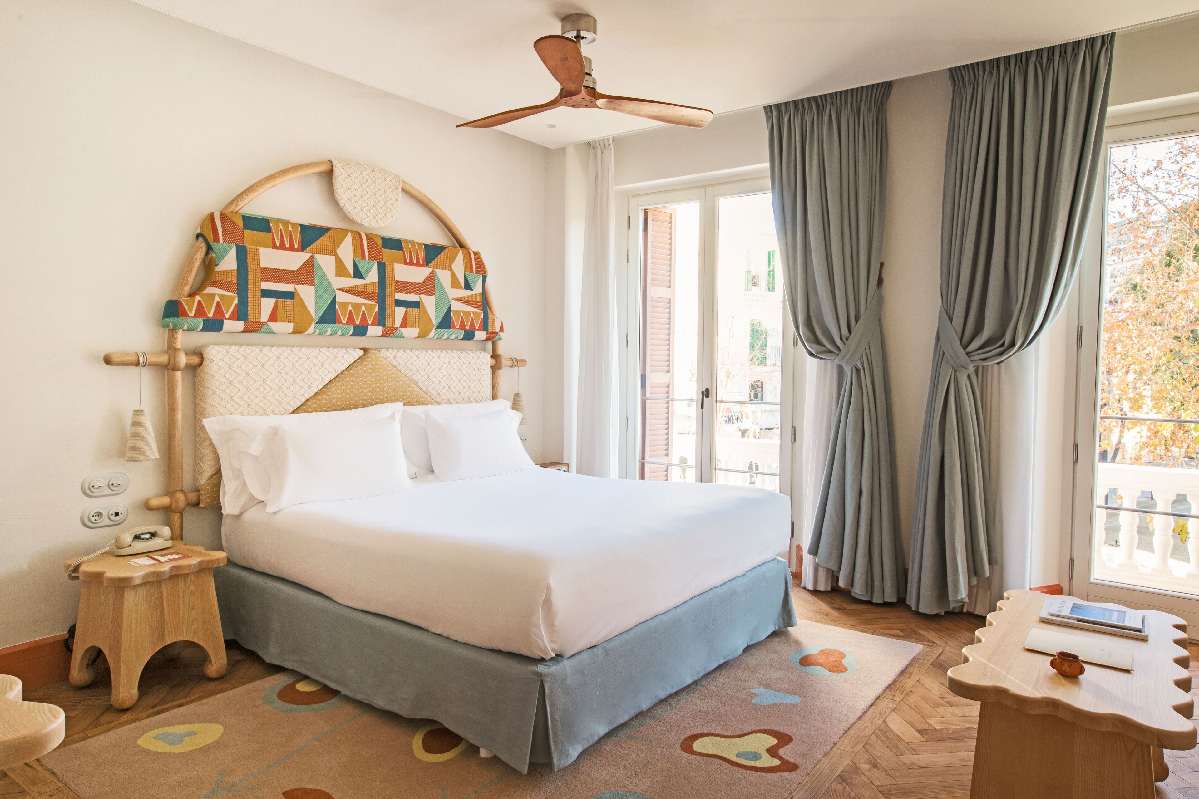 Ein ordentlich eingerichtetes Schlafzimmer mit einem Bett, einem Nachttisch, einem Deckenventilator und Blick ins Freie durch ein Fenster mit Vorhängen.