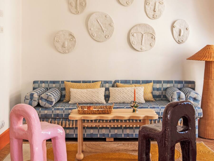 A living room with a patterned sofa, a coffee table, and two chairs. There are wall clocks and a lamp on the wall.