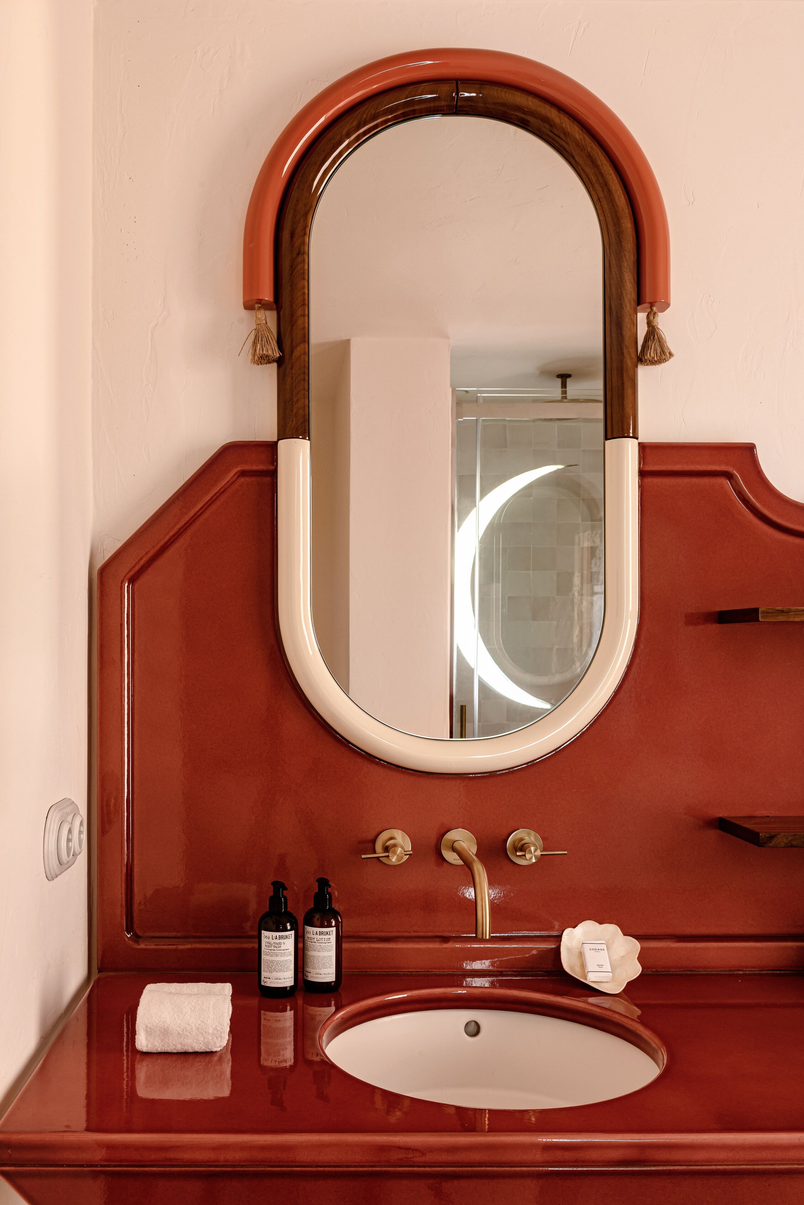 A bathroom with a wooden vanity, a round mirror with a tassel, a sink, and two bottles of lotion.