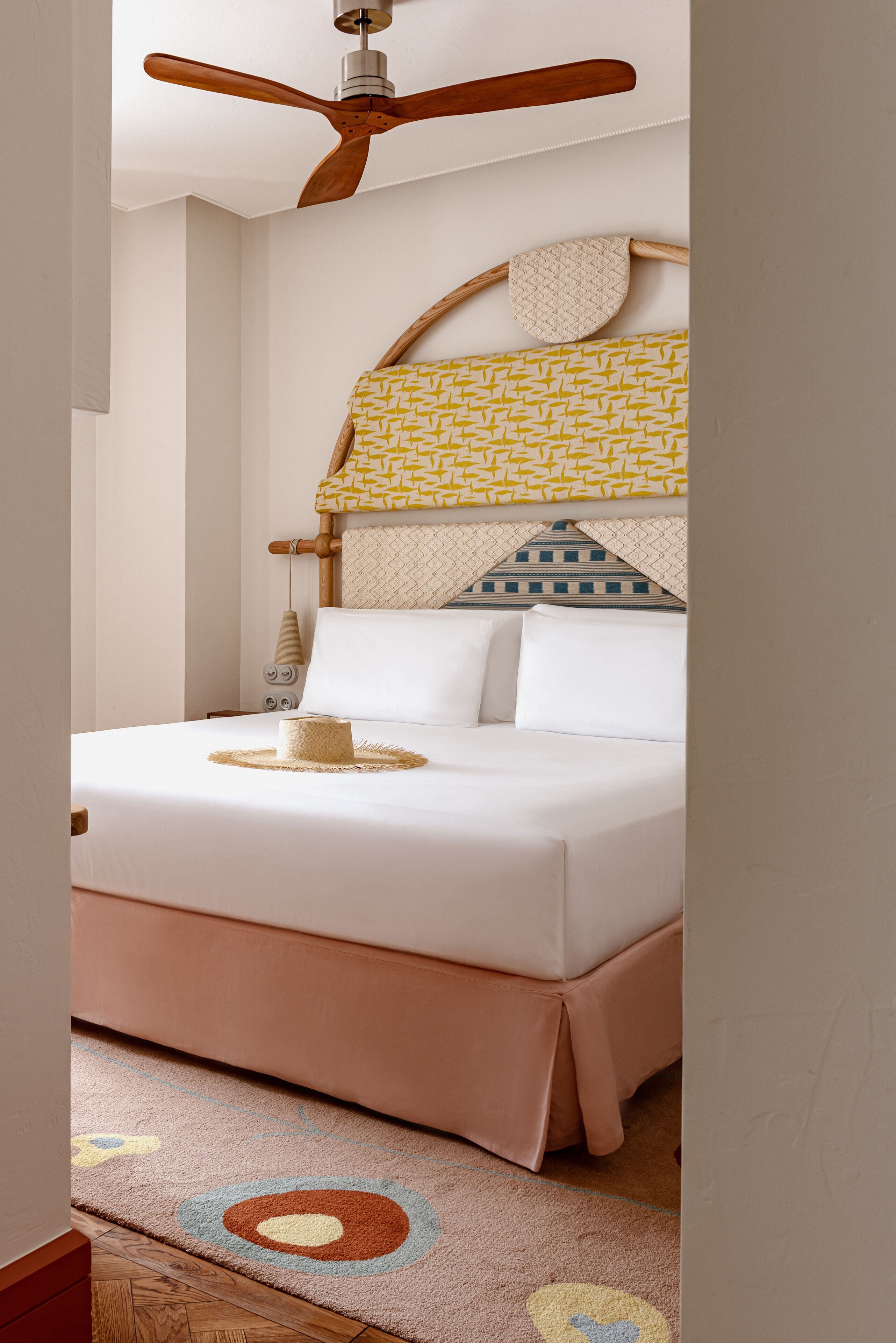 Un dormitorio con cama, ventilador de techo, cabecero estampado y lámpara de noche.