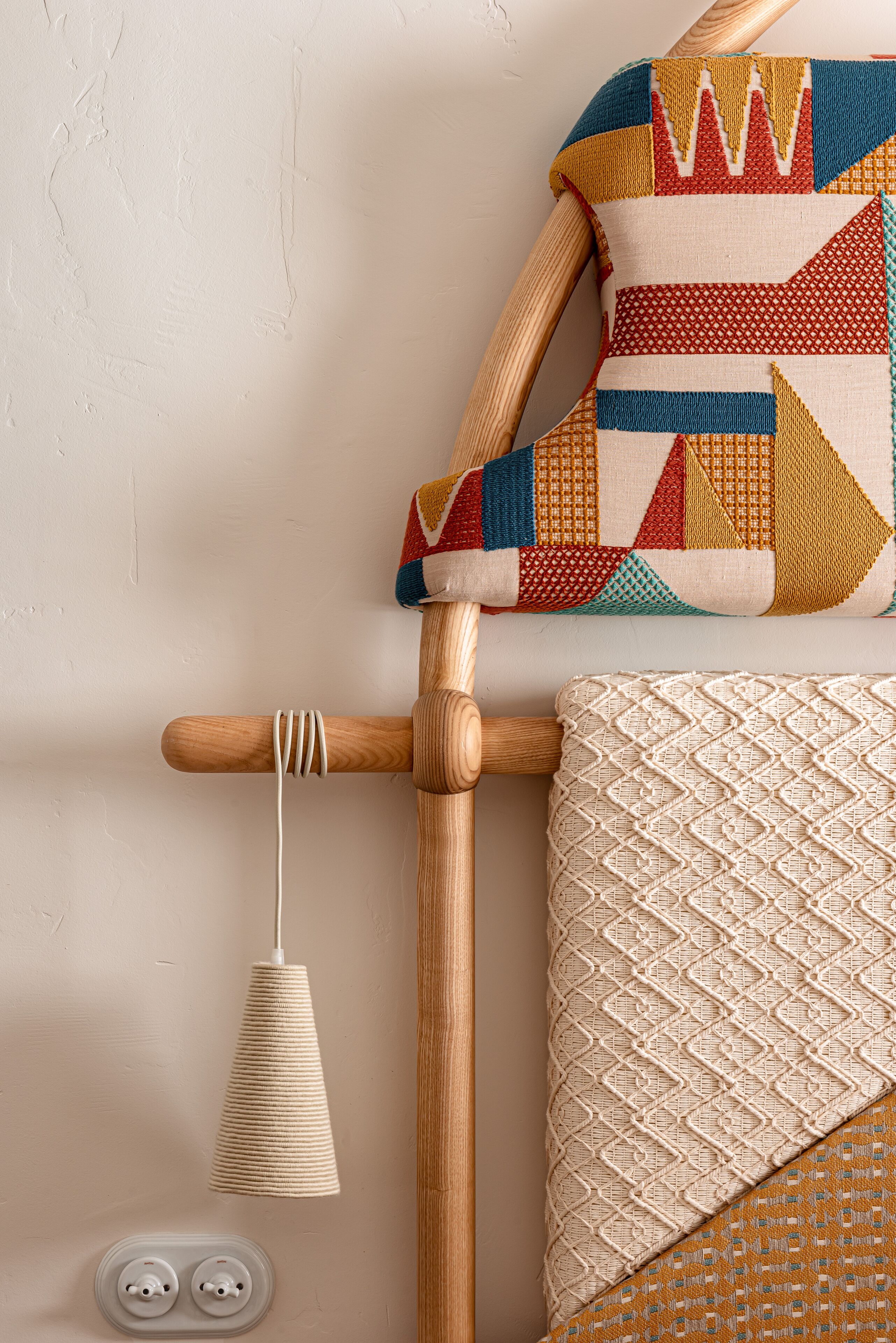 Una silla de madera con asiento y respaldo de mimbre, un cojín colorido y una lámpara fijada a la pared.