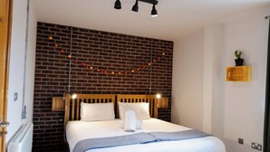 Un lit bien fait, avec une tête de lit en bois, un oreiller blanc et une serviette soigneusement pliée. Une guirlande de petites lumières orange et jaunes est accrochée au mur de briques derrière le lit.