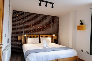 Una cama bien hecha con cabecera de madera, almohada blanca y toalla doblada cuidadosamente. Una guirnalda de luces anaranjadas y amarillas adorna la pared de ladrillo detrás de la cama.