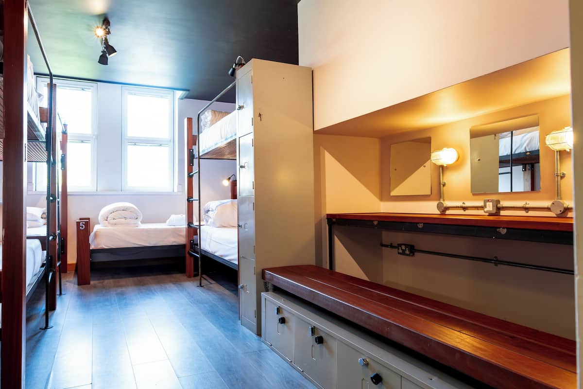 Habitación de hotel pequeña con una litera, un banco de madera, un escritorio, un espejo y una ventana.
