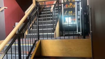 Una escalera con barandilla de madera y balaustrada metálica que conduce a un área con máquinas expendedoras de aperitivos y bebidas.