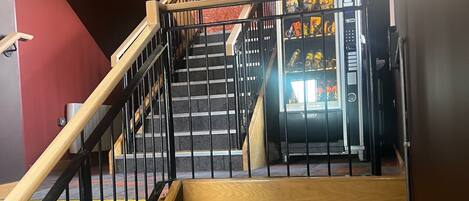 Una escalera con barandilla de madera y balaustrada metálica que conduce a un área con máquinas expendedoras de aperitivos y bebidas.