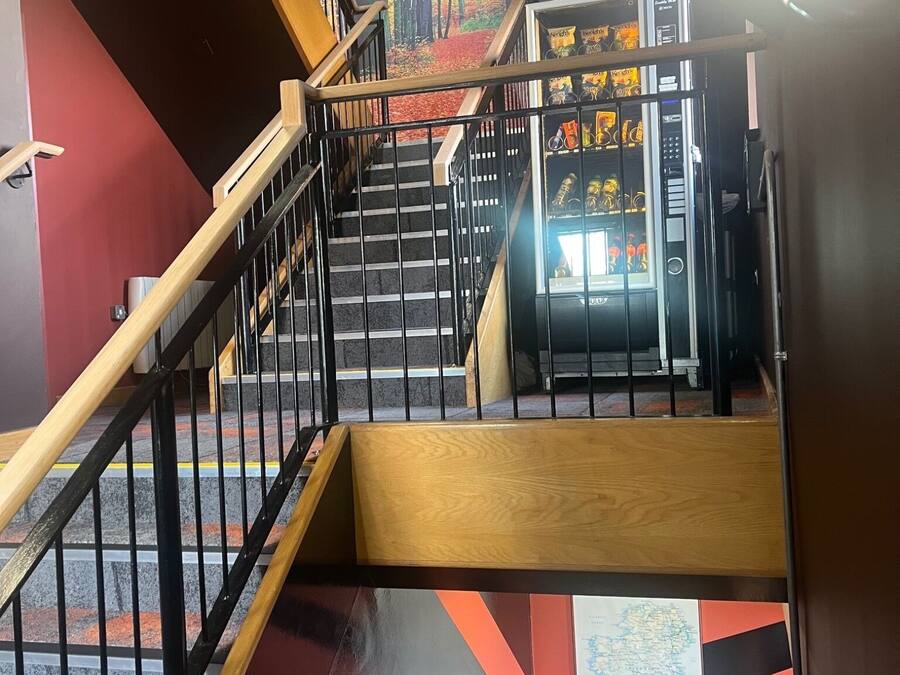 Una escalera con barandilla de madera y balaustrada metálica que conduce a un área con máquinas expendedoras de aperitivos y bebidas.
