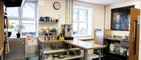Una cocina con paredes de baldosas blancas, un reloj, una cafetera y una ventana con vistas a árboles.
