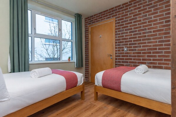 Een kamer met twee houten bedden, een bakstenen muur, een raam met gordijnen en een deur.