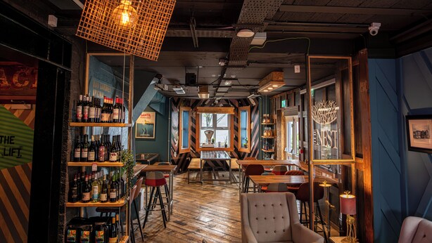 Un restaurant chaleureux avec un plancher en bois, un coin bar où sont exposées des bouteilles de vin, et une table centrale entourée de chaises.