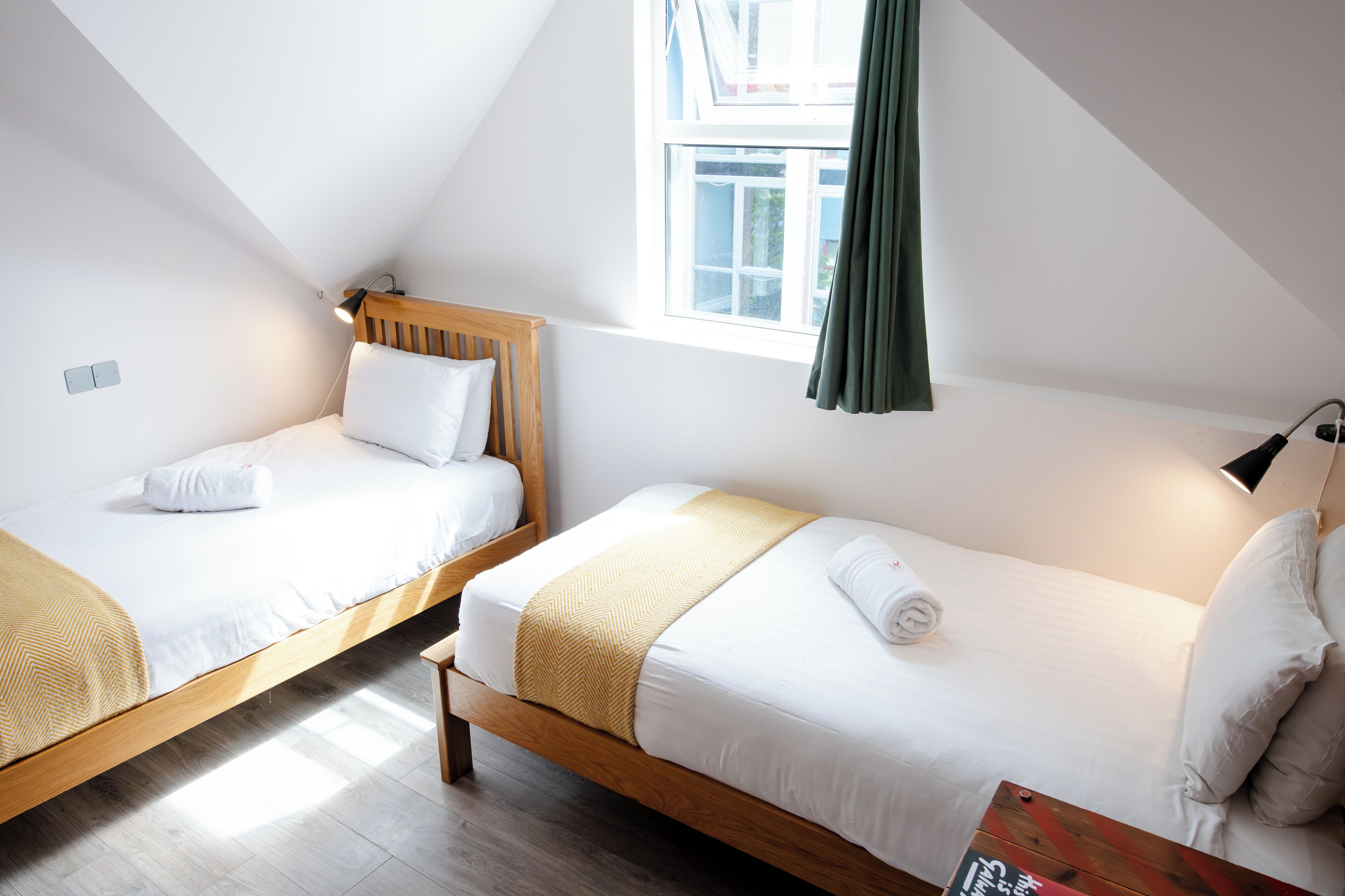 A room with two single beds, wooden headboards, white bedding, a wooden bench, a table with a magazine, and a window with a curtain.