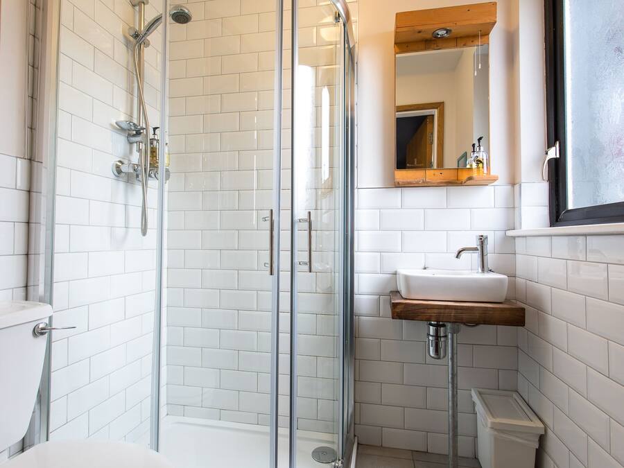 Une salle de bain moderne avec une douche à parois de verre, des murs recouverts de carreaux blancs, un lavabo en bois et un miroir au-dessus du lavabo.