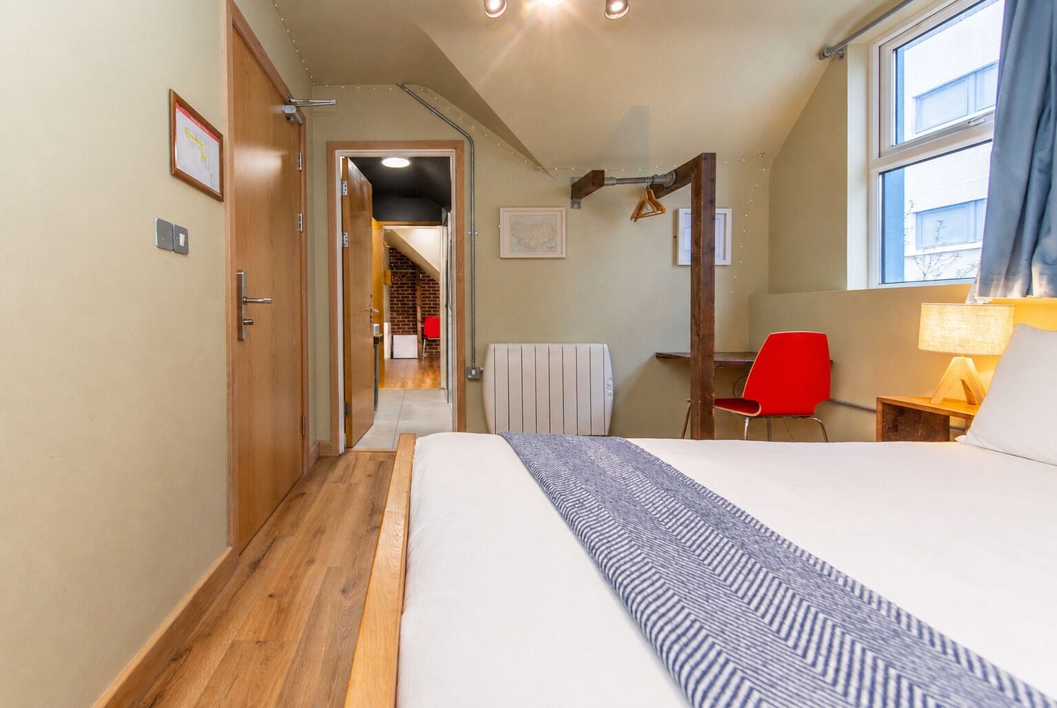 A hotel room with a bed, a red chair, a desk, a window, and a view of a staircase leading to another room.