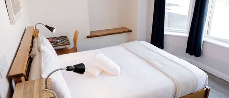A neatly made bed with white linens, a bedside lamp, a wooden nightstand, and a small wooden shelf above the bed.