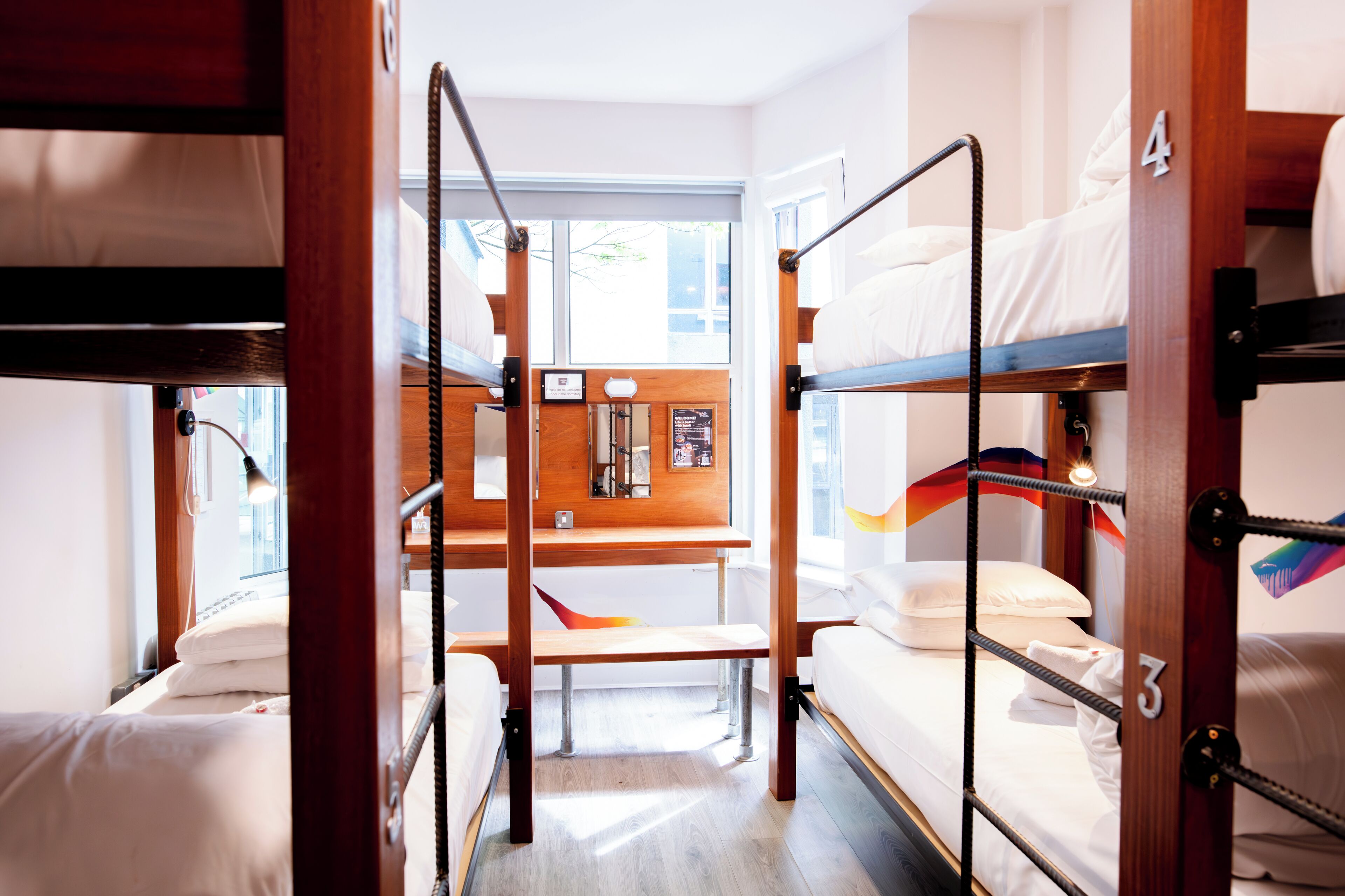 Une chambre de type dortoir, avec des lits superposés, un bureau en bois et une fenêtre équipée de stores.