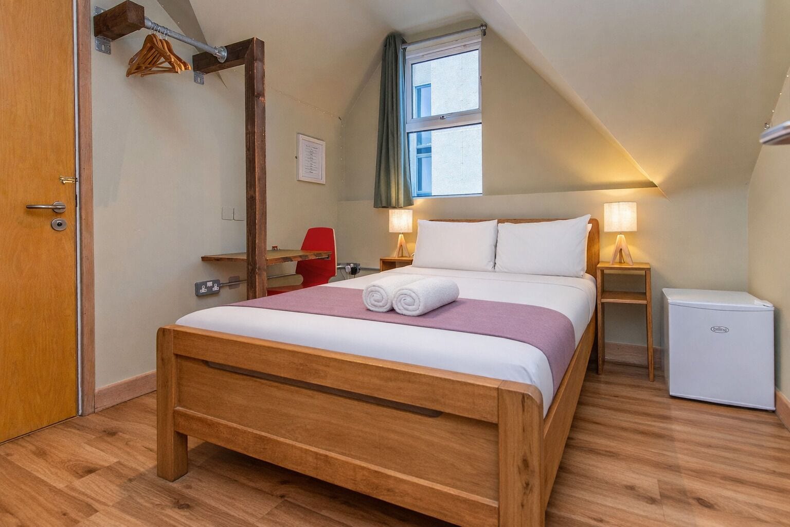 Een slaapkamer met een houten bed, een bureau, een rode stoel, een kleine koelkast en een raam met gordijnen.