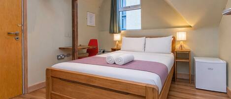 Een slaapkamer met een houten bed, een bureau, een rode stoel, een kleine koelkast en een raam met gordijnen.