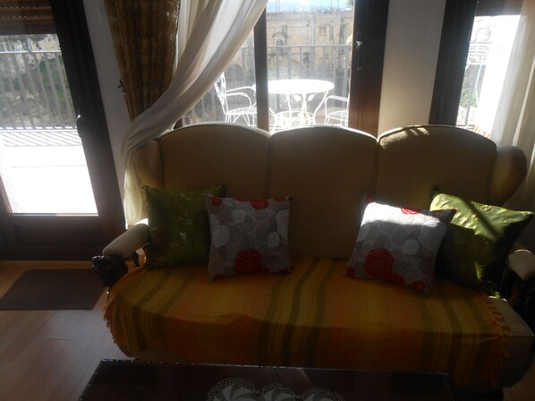 Un sofá con cojines estampados, suelo de madera y vistas a muebles de exterior a través de una ventana.