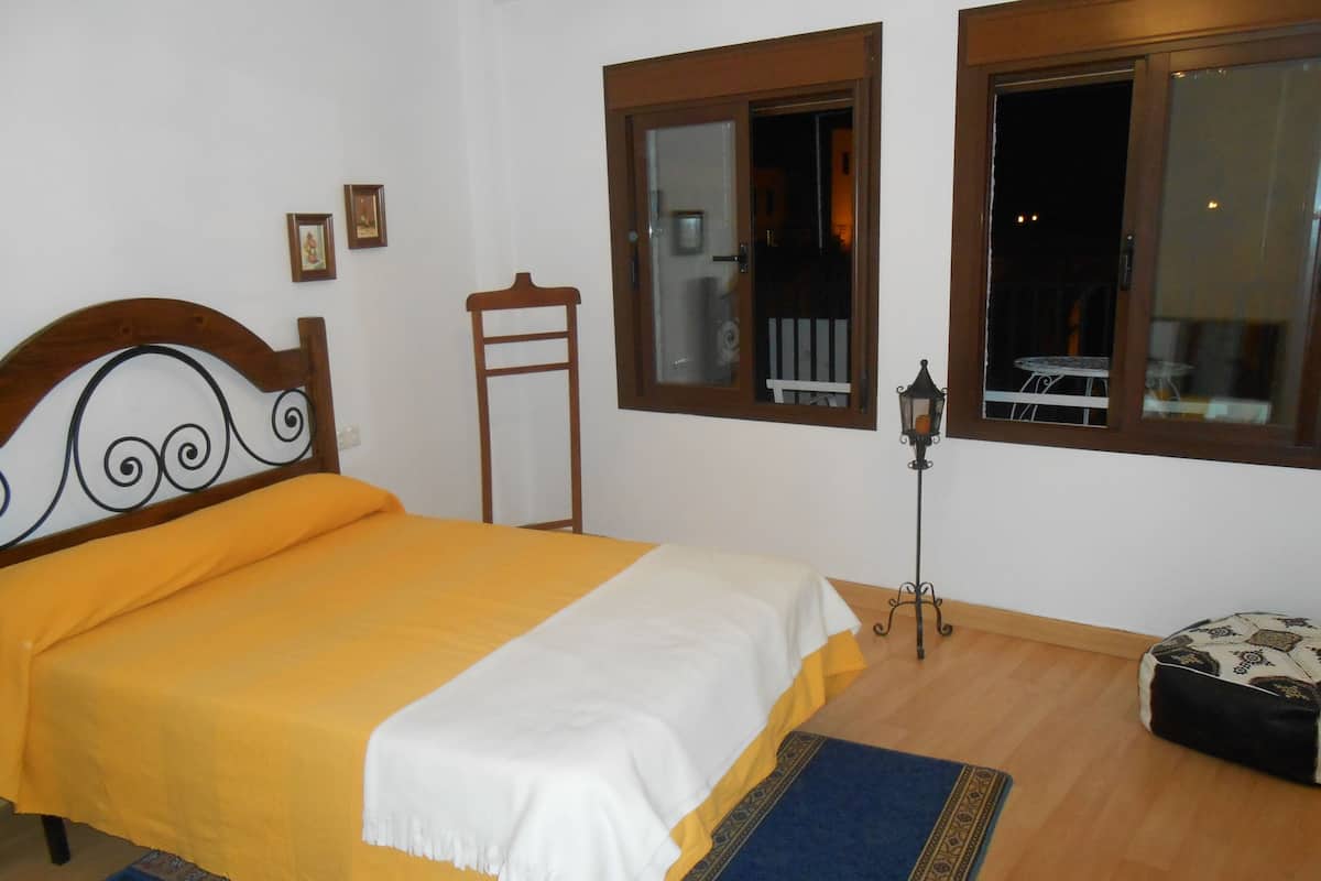 Een slaapkamer met een bed, een houten hoofdbord, een nachtkastje, een lamp, een raam en een kleine tafel.